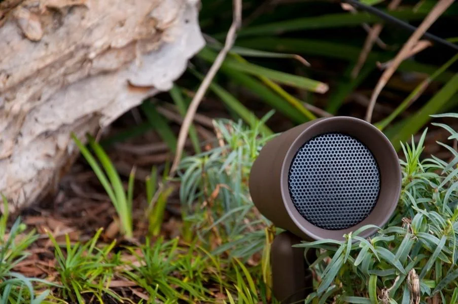 Bronze landscape speaker staked low in a planting bed beside a weathered rock.